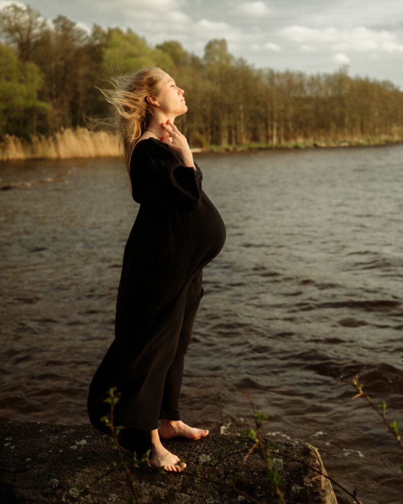 Gravid kvinna i lång svart klänning står vid strandkanten vid Aspen i Lerum.