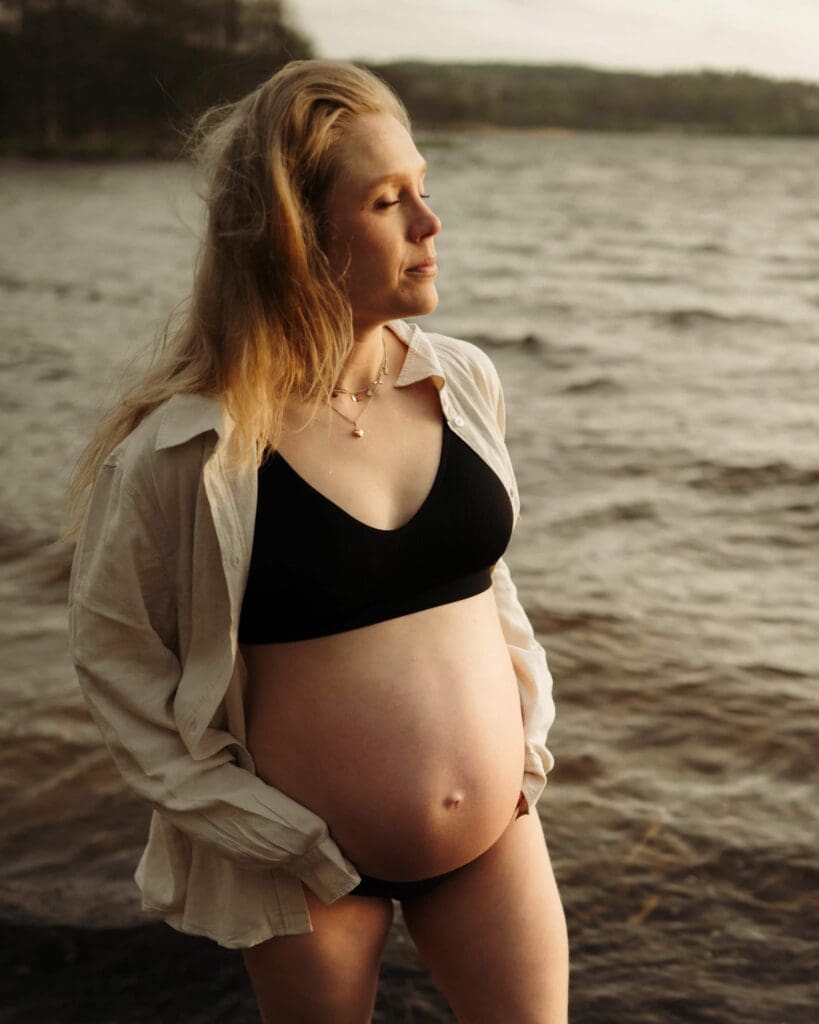Gravid kvinna med svarta underkläder och en linneskjorta över axlarna står vid vattnet vid Aspen i Lerum.