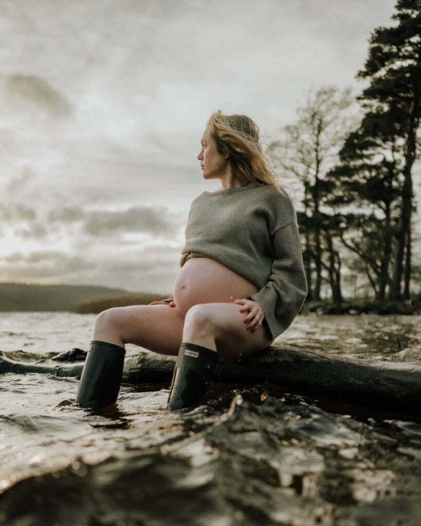Gravid kvinna med stickad tröja uppdragen över magen sitter på en gren vid vattnet i Apen i Lerum.