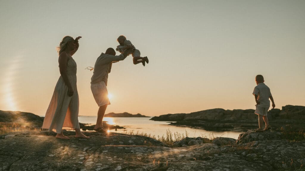 Familj som står på en klippa i solnedgången på Öckerö där pappan håller ett barn och tappar sina solglasögon.