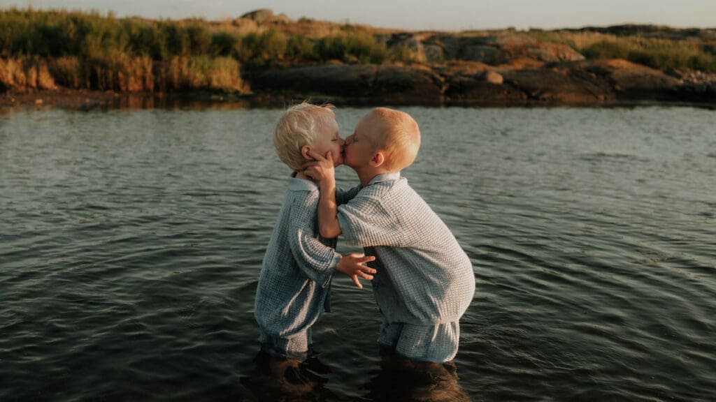 Två yngre barn står i havet på Öckerö och pussar varandra i solnedgångsljuset.