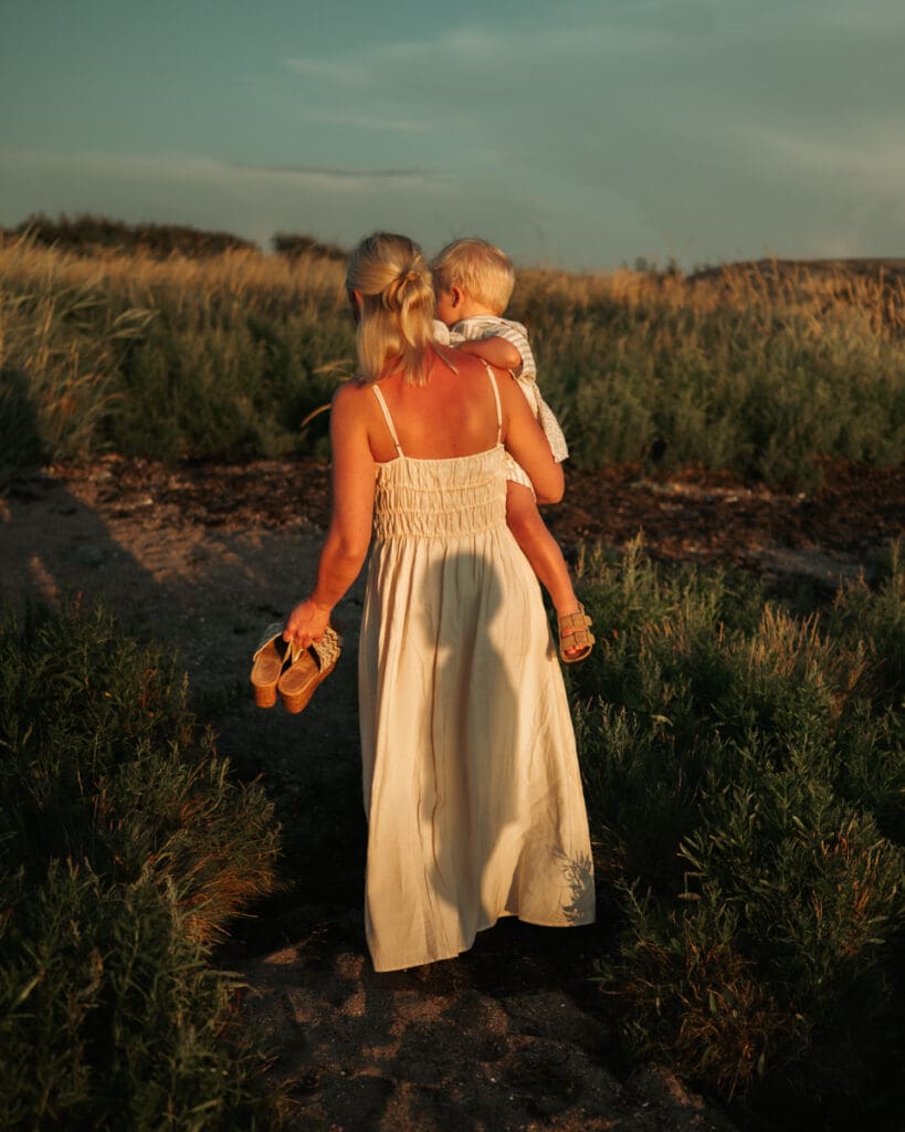 Bild bakifrån på en mamma i skir klänning som håller sitt ena barn på armen och skor i andra handen med siluetten av hennes andra barn på klänningen.