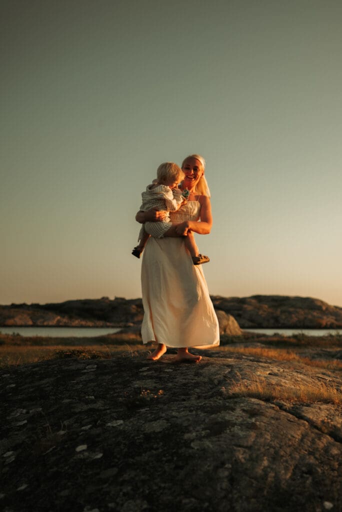 Mamma dansar på klipporna vid havet på Öckerö i solnedgången med sin lilla son i famnen.