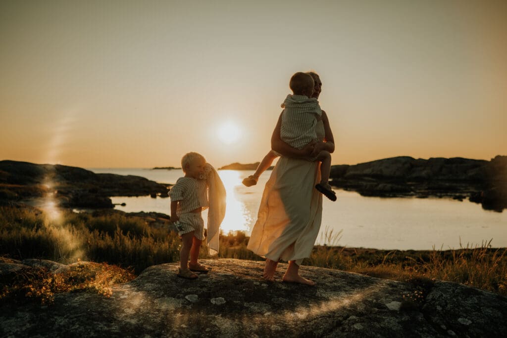 Mamma dansar med sin ena son i famnen och den andra står bredvid och tittar på vid solnedgången på en klippa på Öckerö.