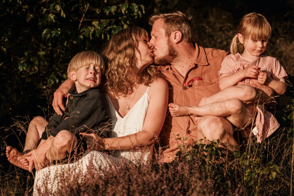 Mamma och pappa som sitter i gräset och pussar varandra på en äng i Lerum med vars ett barn i knäet, sonen gör en rolig grimas.